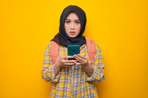 Verwarde jonge Aziatische moslimvrouw student in Plaid shirt en rugzak met behulp van mobiele telefoon geïsoleerd op gele achtergrond Onderwijs school universiteit college concept