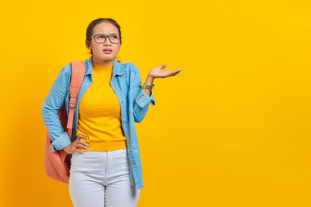 Verwarde jonge Aziatische vrouw student in denim outfit met rugzak nee zeggen met handpalmen en opzij kijken geïsoleerd op gele achtergrond Onderwijs in universiteitscollege concept