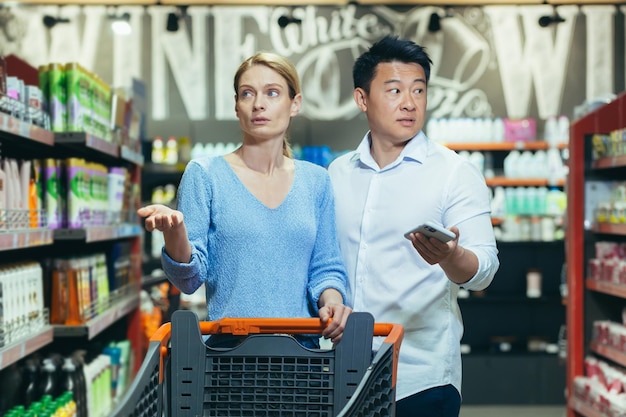 Verwarde jonge internationale koppelvrouw en aziatische man die tussen rijen loopt met een boodschappenwagentje en