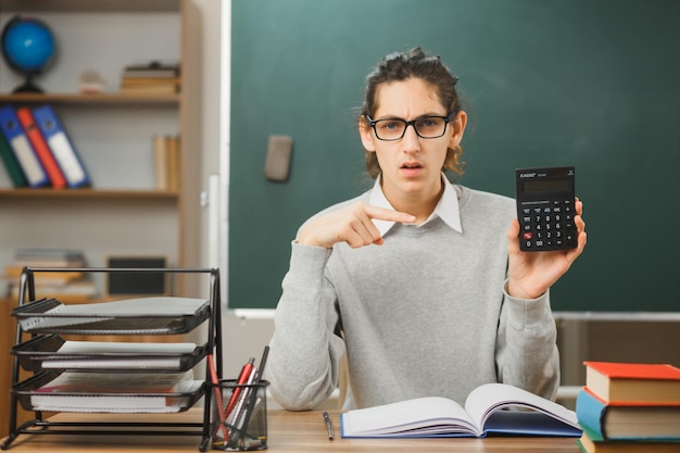 verwarde jonge mannelijke leraar die vasthoudt en wijst naar de rekenmachine die aan het bureau zit met schoolhulpmiddelen aan in de klas