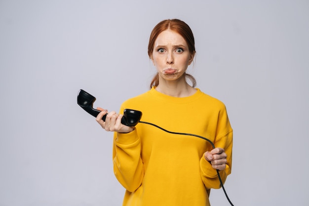 Verwarde jonge vrouw in gele trui met handset ontvanger van retro telefoon en camera kijken