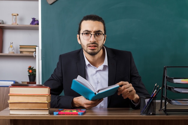 Verwarde mannelijke leraar met een bril die een boek aan tafel vasthoudt met schoolhulpmiddelen in de klas