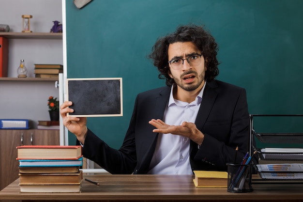 Verwarde mannelijke leraar met een bril die vasthoudt en wijst met de hand naar een mini-bord aan tafel met schoolhulpmiddelen in de klas