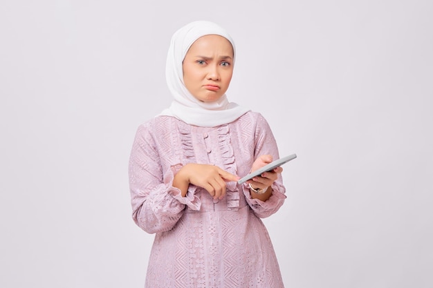 Verwarde mooie jonge Aziatische moslimvrouw die hijab en paarse jurk draagt die mobiele telefoon vasthoudt en naar de camera kijkt die op een witte studioachtergrond wordt geïsoleerd