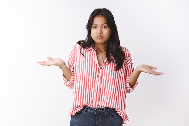 Verwarde onzekere vrouwelijke newbie die geen idee heeft en haar schouders ophaalt en de handen zijwaarts spreidt in ontzetting en frustratie, niet op de hoogte en onzeker hoe de fout is gebeurd tegen de witte muur