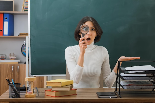 verwarde spreidende handen jonge vrouwelijke leraar kijkend naar camera met vergrootglas zittend aan bureau met schoolhulpmiddelen in de klas