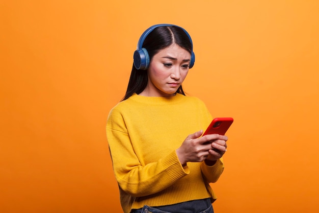 Verwarde verbaasde vrouw die zich hulpeloos voelt vanwege een defect smartphoneapparaat. Twijfelachtige jonge volwassen aziatische persoon die een koptelefoon draagt terwijl hij een mobiele telefoon op een oranje achtergrond heeft.