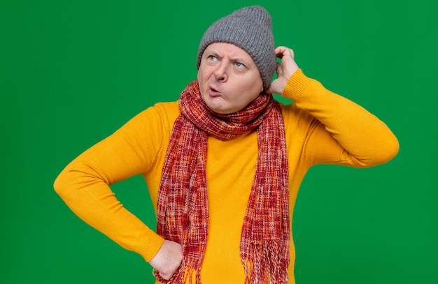 Foto verwarde volwassen man met wintermuts en sjaal om zijn nek die zijn hoofd krabt en naar de zijkant kijkt