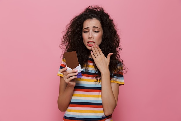 verwarde vrouw 20s met krullend haar met chocoladereep geïsoleerd op roze