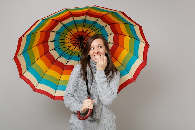 Verwarde vrouw in grijze trui, sjaal opzoeken, nagels knagen, kleurrijke paraplu geïsoleerd op grijze achtergrond houden. Gezonde mode levensstijl mensen emoties, koude seizoen concept. Bespotten kopie ruimte.