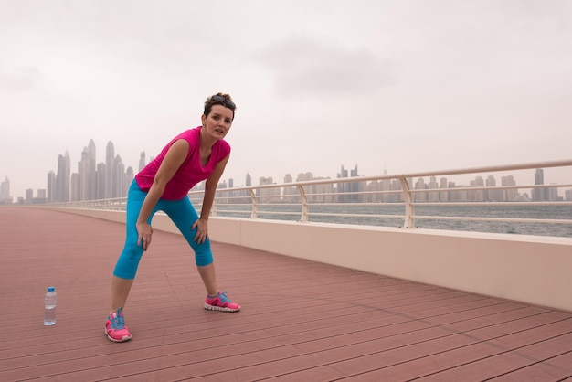 very active young beautiful woman stretching and warming up on the promenade along the ocean side with a big modern city in the background to keep up her fitness levels as much as possible