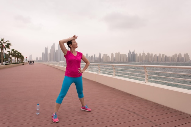 Very active young beautiful woman stretching and warming up on\
the promenade along the ocean side with a big modern city in the\
background to keep up her fitness levels as much as possible