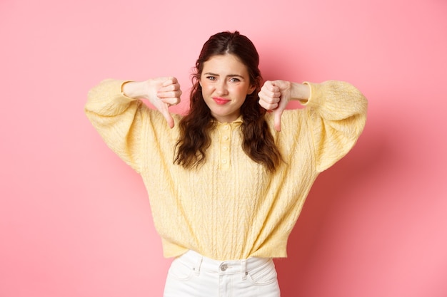 Very bad. Disappointed young woman complaining on something awful, showing dislike with thumbs down, grimacing and frowning, shaking head, give negative feedback, pink wall