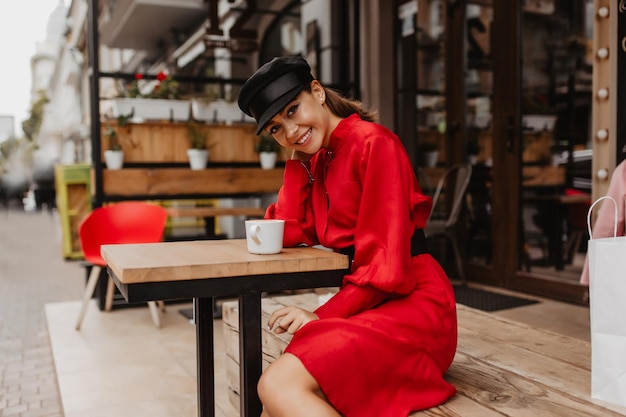 Very beautiful and tender girl of 20 years old in elegant dress and fashionable hat is resting on terrace of  cafe enjoying aroma of coffee