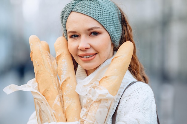 A very beautiful woman in a blue knit band and a white sweater advertises bakery products. He holds packages of loaves in his hands. Blurry outdoor background. Marketing portrait of bakery.