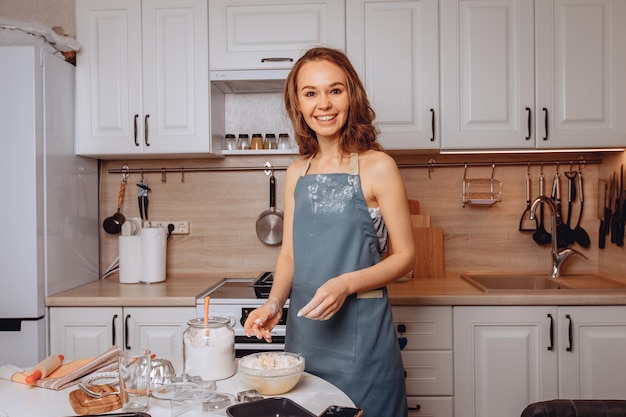 Very cute young girl in blue apron is standing in kitchen of the house and preparing biscuits. Kneading dough with her hands. Hands and apron are slightly stained with flour due to cooking process.