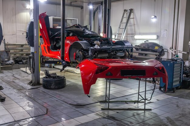 Very expensive and powerful sports car of red color disassembled for repair lifted on a lift in auto service next to it is a removed bumper