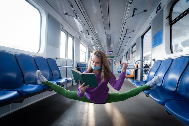 매우 유연한 마스크를 착용한 여성이 체조 분할에 앉아 지하철에서 책을 읽고 있습니다. 건강한 생활 방식, 유연성 및 요가의 개념