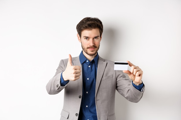 Photo very good bank. businessman in suit show plastic credit card and thumb-up, recommending, standing on white background.