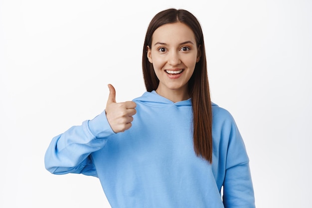 Very good. Image of young woman in hoodie showing thumb up in approval, like and agree, praise good work, satisfied with quality, recommend smth, white background.