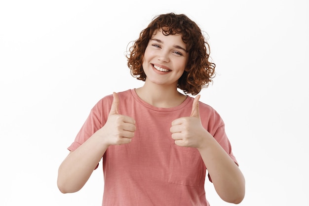 Very good, nice job. Smiling young curvy girl showing thumbs up, give positive feedback, recommend something nice, praising great work, standing on white