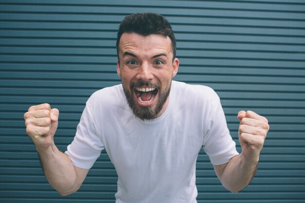 Very happy man is standing and looking at camera. He is holding hands in fists and opening mouth very wide. Isolated on striped 