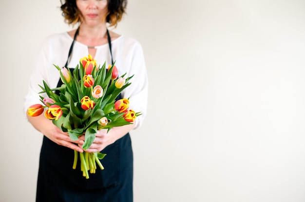 Donna fiorista molto bella che tiene un mazzo di bellissimi fiori sboccianti colorati di tulipani freschi sullo sfondo della parete.