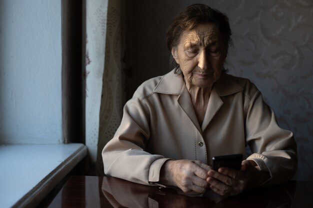 A very old senior Caucasian grandmother with deep wrinkles sits home, uses her smart phone on outstretched arm front camera for a video call.