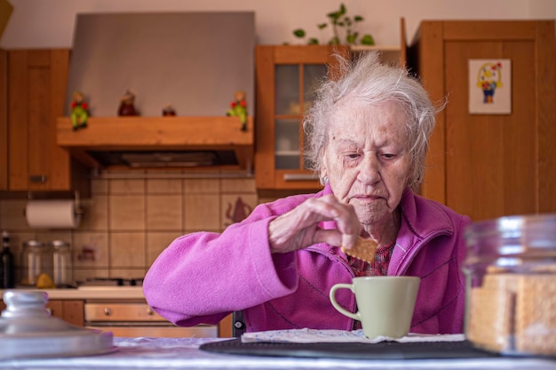 very old woman alone in her kitchen dips cookie in cup of latte coffee during breakfast