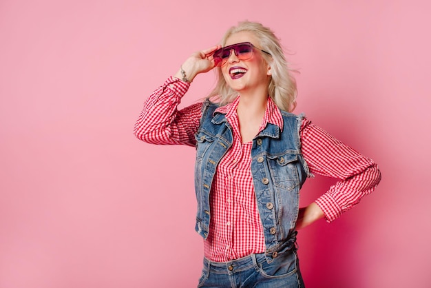 Very positive and happy adult blond woman dressed in shirt and blue jeans posing on pink background