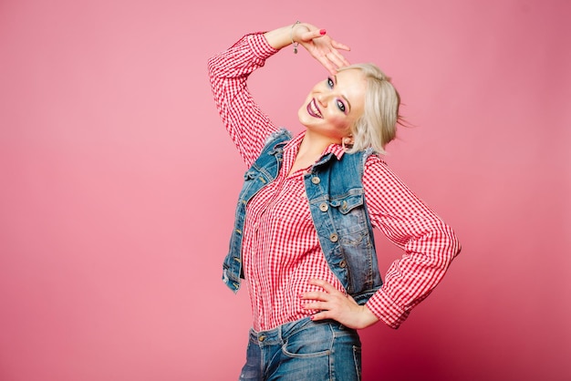 Very positive and happy adult blond woman dressed in shirt and blue jeans posing on pink background