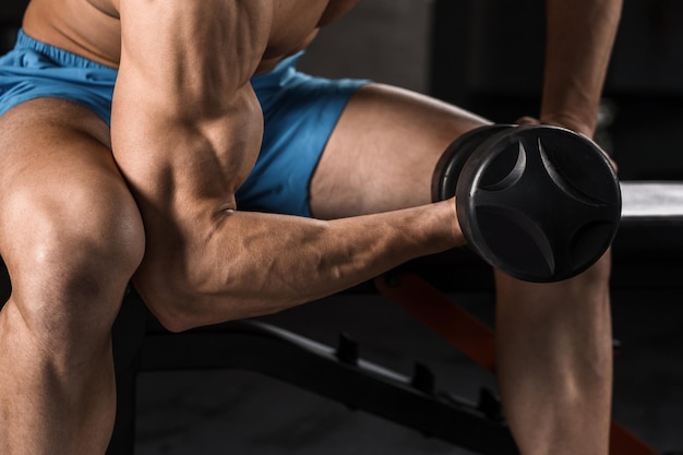 Very power athletic guy bodybuilder execute exercise with dumbbells at dark gym. Muscular man. Part of the body