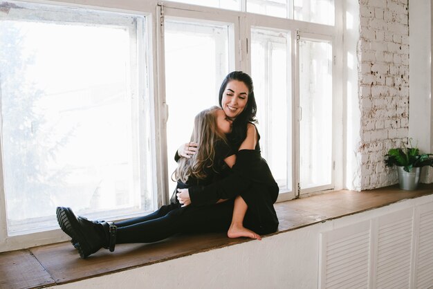 Very stylish mom and daughter with bare feet hug on the windowsill