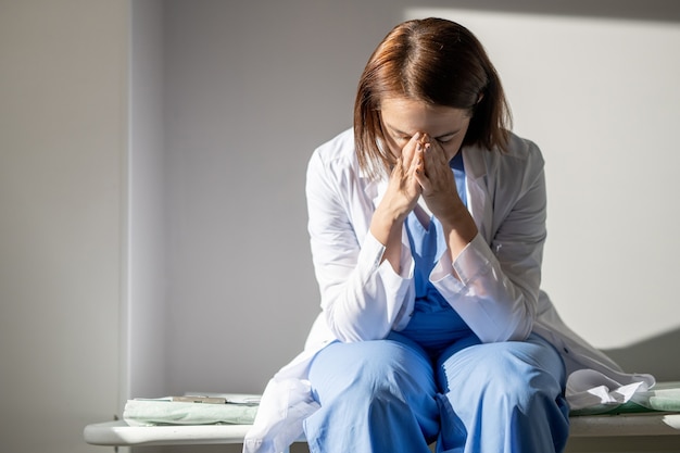 Very tired young female assistant or caregiver of covid hospital sitting on medical cart against wall of chamber and praying for someone