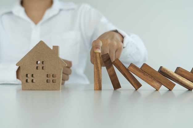 Verzekering met handen beschermt een huis Het houten dominoblok staat op het punt op het huis te vallen Opstalverzekering of huisverzekeringsconcept