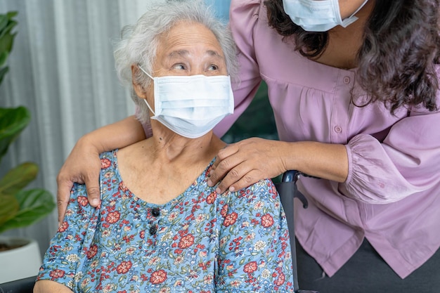 Verzorger helpt aziatische senior of oudere oude dame vrouw zittend op een rolstoel en het dragen van een gezichtsmasker ter bescherming van de veiligheidsinfectie covid 19 coronavirus