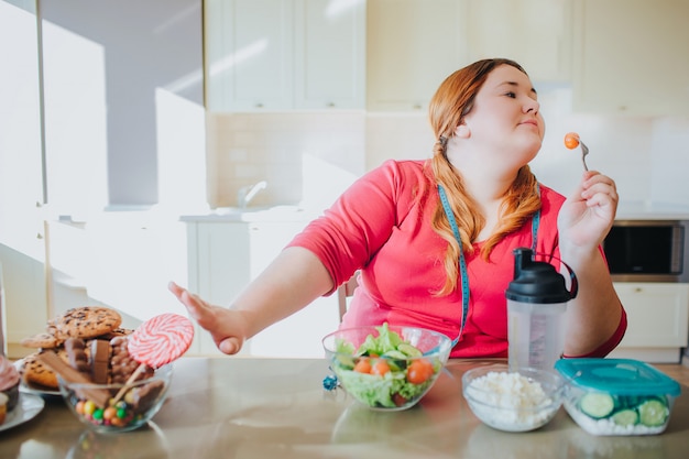 Vette jonge vrouw in keuken die en gezond voedsel zitten eten