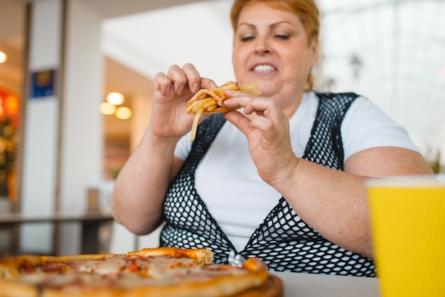 Vette vrouw die pizza met frietjes eet in winkelcentrumrestaurant, ongezond voedsel Vrouwelijke persoon met overgewicht aan tafel met junk-diner, obesitasprobleem