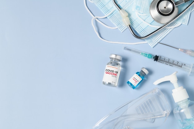 파란색 배경에 수술 마스크에 주사기와 청진기가 달린 Covid 19 백신의 바이알, 코로나 바이러스 또는 COVID-19 발발 개념 예방