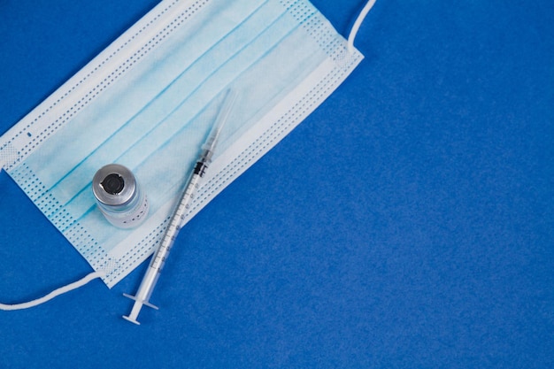 Vial of Covid19 vaccine a syringe for injection and a blue mask on a blue background SARSCoV2 vaccination treatment Top view copy space