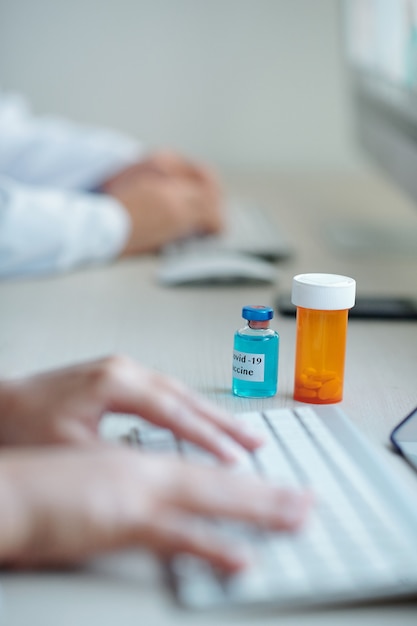 Vial of vaccine against coronavirus and bottle of pills on desk in front of doctor working on laptop