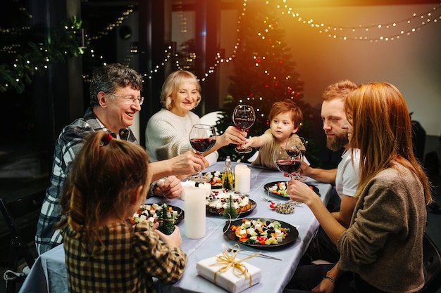 Vibrant big family on christmas gathering. Sitting around a big dining table, clinking wine glasses, celebrating. Christmas tree and garlands in background.