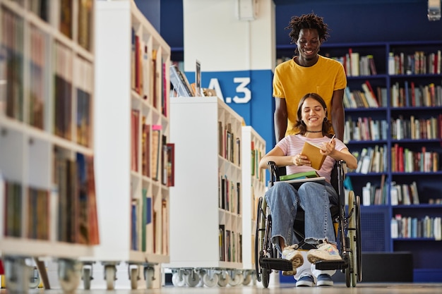 Foto vibrante scatto a figura intera di un giovane nero che assiste una studentessa con disabilità in biblioteca e