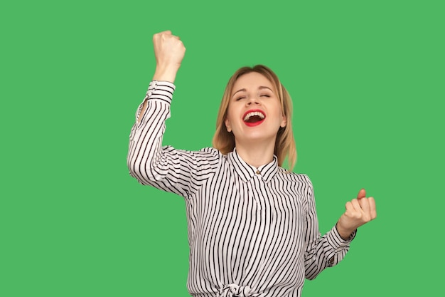 Victory celebration. Excited blond girl screaming loudly and raising hands in gesture I did it, enjoying success with ecstatic satisfied expression. indoor studio shot isolated on green background