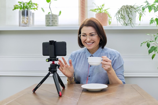 Video blog vlog stream vrouw kijken naar smartphone webcam op statief video opnemen online praten thuis aan tafel zitten educatief kanaal journalist psycholoog counselor