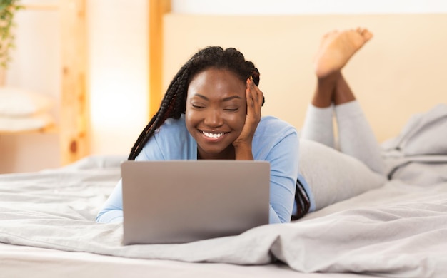 Video Call. Cheerful African American Student Girl Using Laptop Communicating With Family At A Distance Lying In Bed At Home