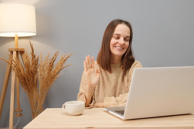 Video call communication Remote employment Friendly brown haired woman wearing beige sweater using computer waving hand greeting hello gesture while sitting at table in home office