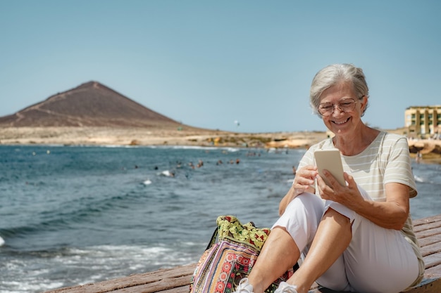 Video call concept Smiling senior woman using phone for online communication sitting at the beach