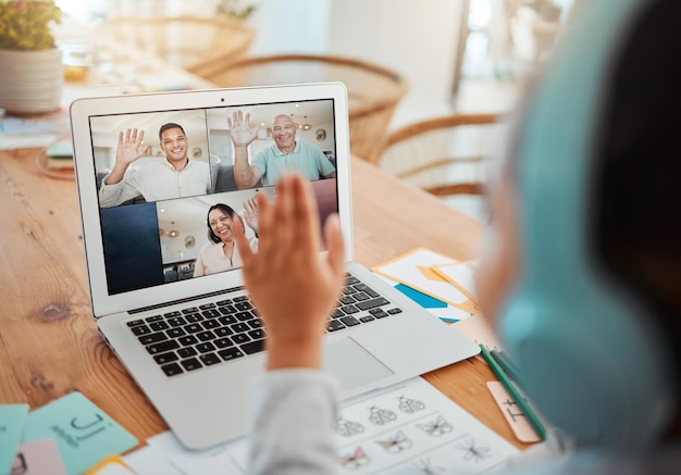 Photo video call laptop and child greeting her family while sitting by the dining room table in her home technology waving and girl kid on a virtual call with her parents and grandfather on a computer