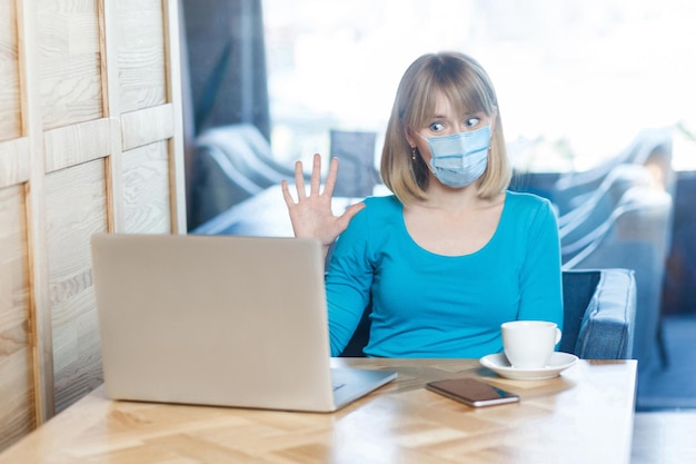 Video call, online working at home. Portrait of young woman with surgical medical mask is sitting and working on laptop and greeting on video call conference. Indoor, medicine and health care concept